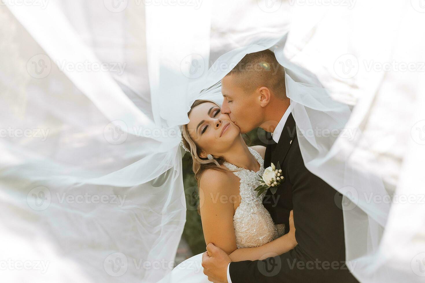 gorgeous happy luxurious brunette bride and elegant stylish groom stand under a veil and embrace tenderly photo