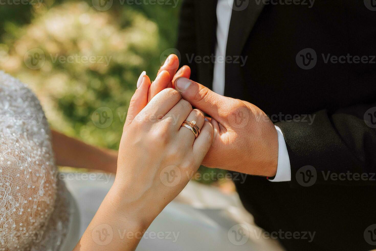 A stylish groom in a black suit and a cute bride in a white dress with a long veil are hugging in the park. Close-up wedding portrait of the hands of the happy newlyweds. photo