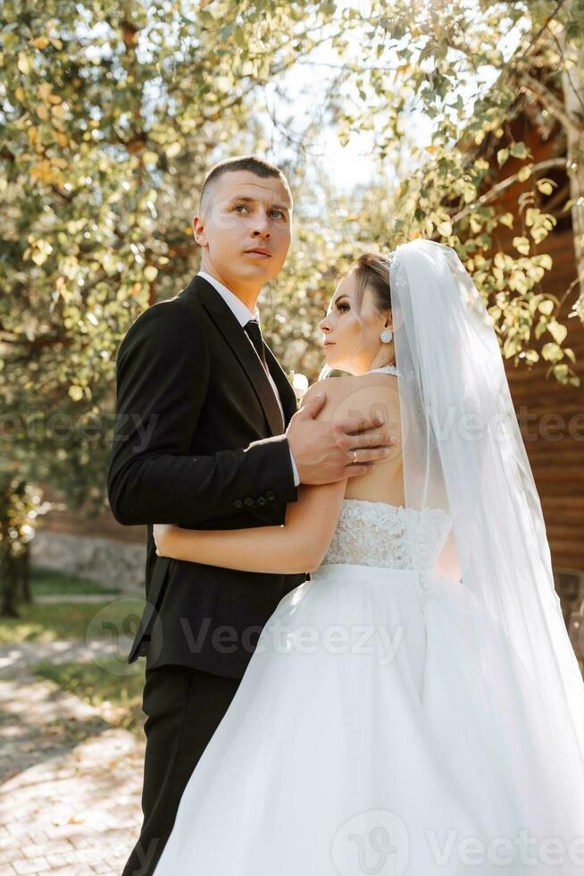 A stylish groom in a black suit and a cute bride in a white dress with a long veil are hugging in a park. Wedding portrait of smiling and happy newlyweds. photo