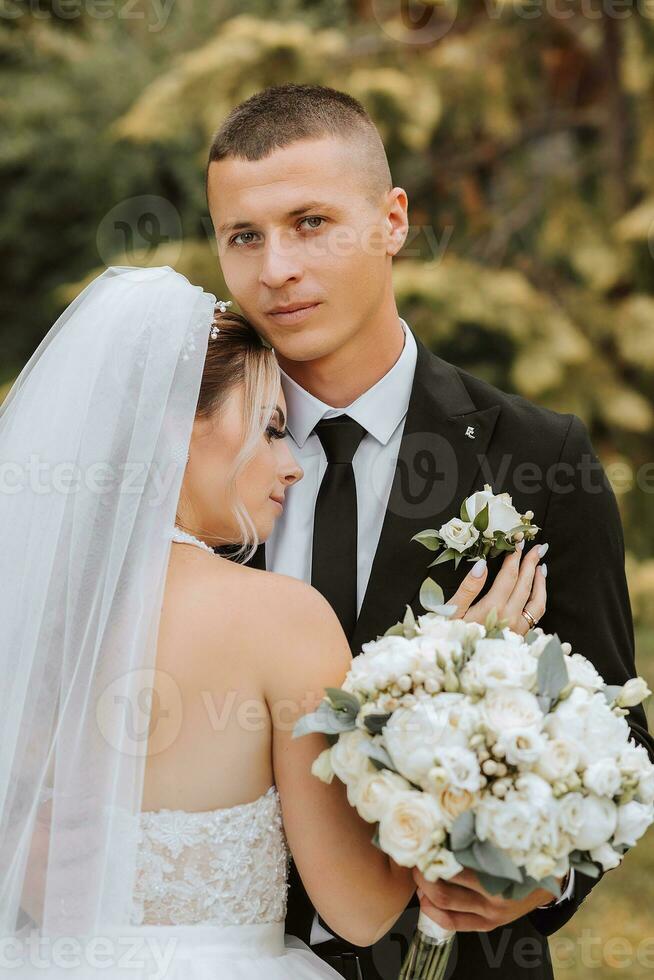 un elegante novio en un negro traje y un linda novia en un blanco vestir con un largo velo son abrazando en un parque. Boda retrato de sonriente y contento recién casados. foto