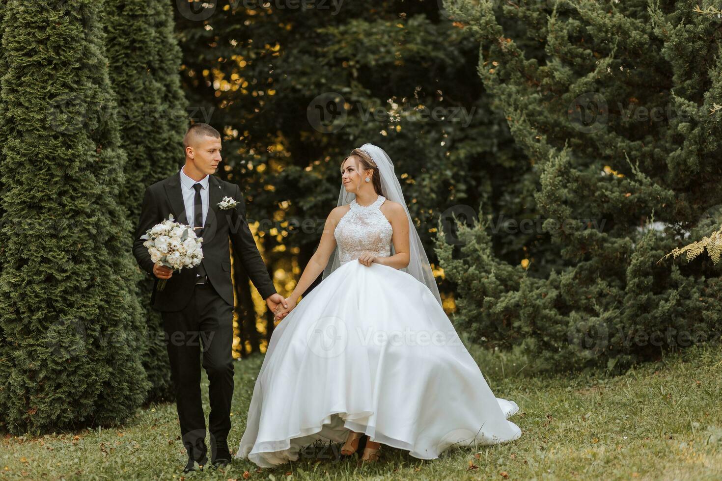 A stylish groom in a black suit and a cute bride in a white dress with a long veil are hugging and walking near green tall trees. Wedding portrait of smiling and happy newlyweds. photo