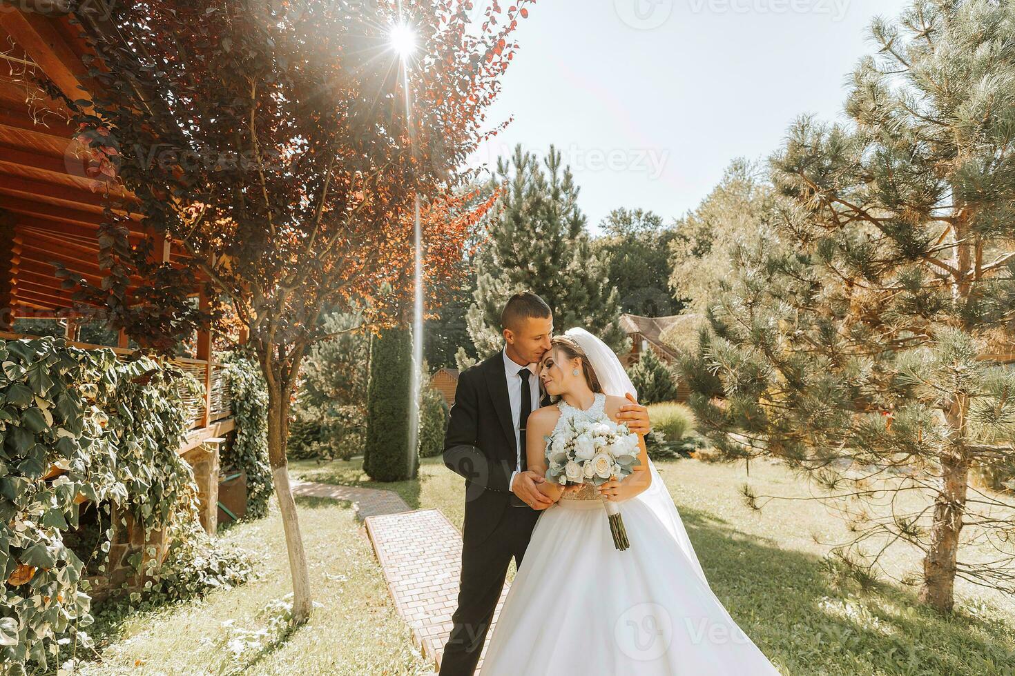 A stylish groom in a black suit and a cute bride in a white dress with a long veil are hugging in a park. Wedding portrait of smiling and happy newlyweds. photo