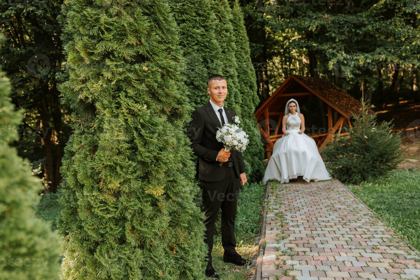 A stylish groom in a black suit and a cute bride in a white dress with a long veil are hugging and walking near green tall trees. Wedding portrait of smiling and happy newlyweds. photo