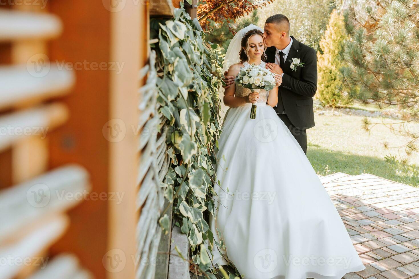 A stylish groom in a black suit and a cute bride in a white dress with a long veil are hugging in a park. Wedding portrait of smiling and happy newlyweds. photo