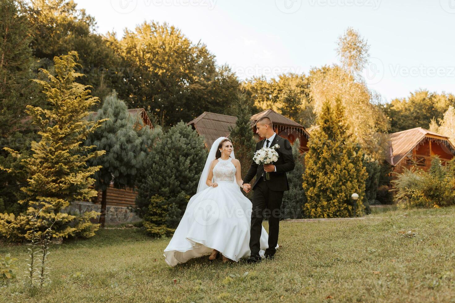 A stylish groom in a black suit and a cute bride in a white dress with a long veil are hugging and walking near green tall trees. Wedding portrait of smiling and happy newlyweds. photo