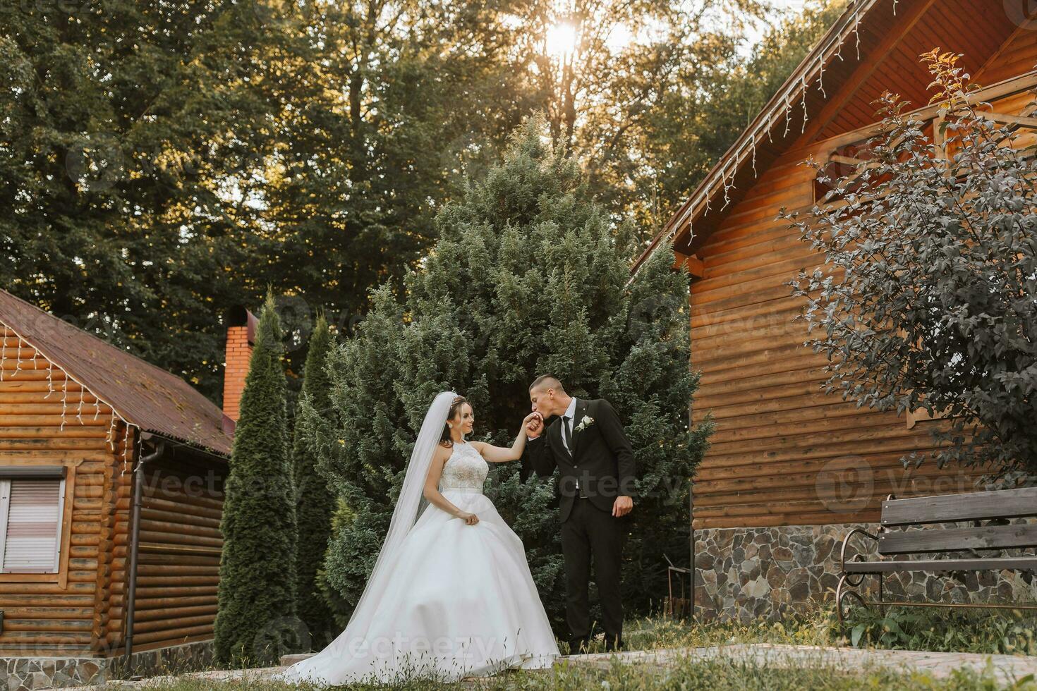 A stylish groom in a black suit and a cute bride in a white dress with a long veil are hugging and walking near green tall trees. Wedding portrait of smiling and happy newlyweds. photo