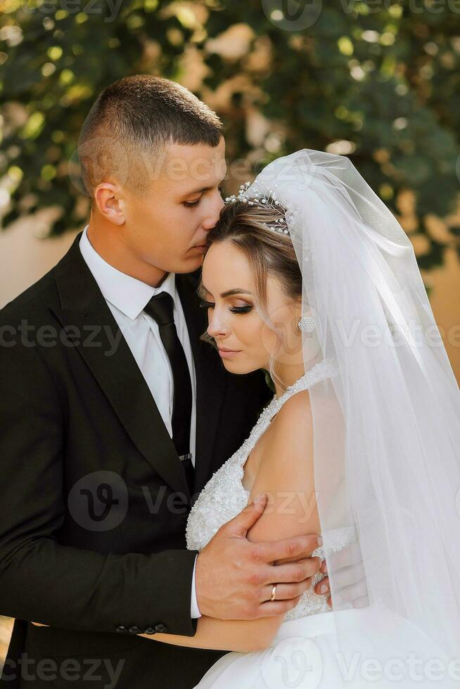 un elegante novio en un negro traje y un linda novia en un blanco vestir con un largo velo son abrazando en un parque. Boda retrato de sonriente y contento recién casados. foto