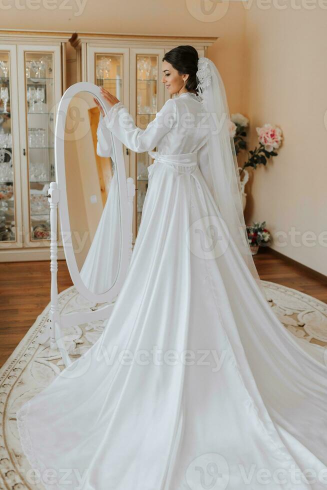 Happy dark-haired brunette bride in a satin robe and professional make-up standing in her room near the mirror and getting ready for her wedding day photo