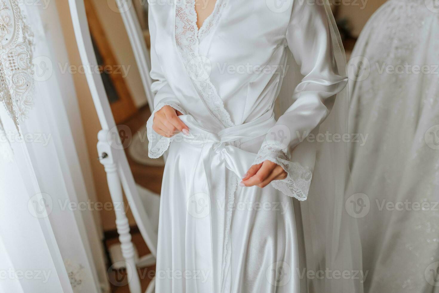Happy dark-haired brunette bride in a satin robe and professional make-up standing in her room near the mirror and getting ready for her wedding day photo