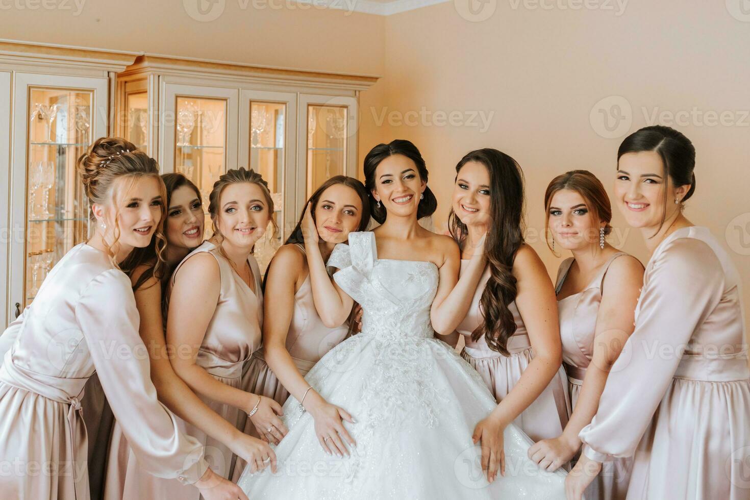 Stylish beautiful bridesmaids in matching silk dresses helping gorgeous brunette bride in white dress get ready for wedding, morning preparations, woman putting on dress photo