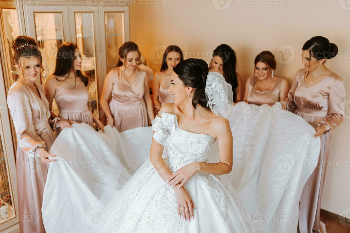 Stylish beautiful bridesmaids in matching silk dresses helping gorgeous brunette bride in white dress get ready for wedding, morning preparations, woman putting on dress photo