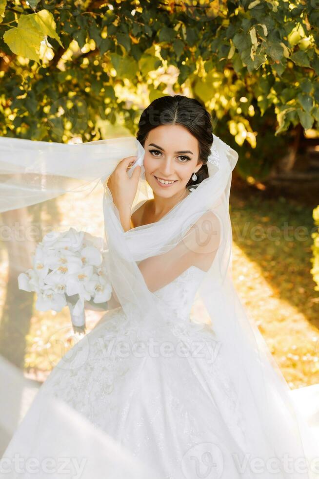 Warm sunny portrait of a happy bride with a bouquet of orchids in her hands, outdoors and at sunset. Warm summer time. A long veil, a luxurious white dress with a train. photo