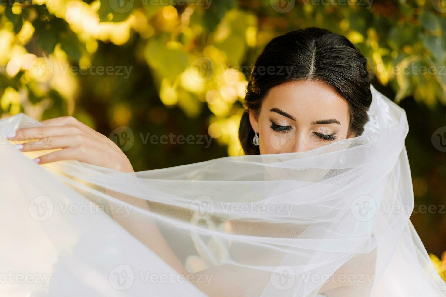 Warm sunny portrait of a happy bride with a bouquet of orchids in her hands, outdoors and at sunset. Warm summer time. A long veil, a luxurious white dress with a train. photo