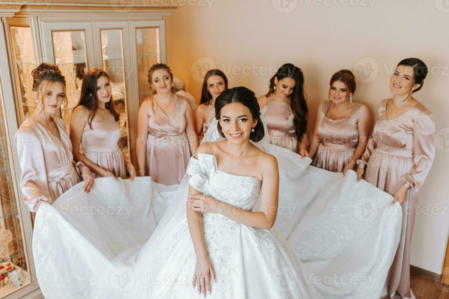 Stylish beautiful bridesmaids in matching silk dresses helping gorgeous brunette bride in white dress get ready for wedding, morning preparations, woman putting on dress photo