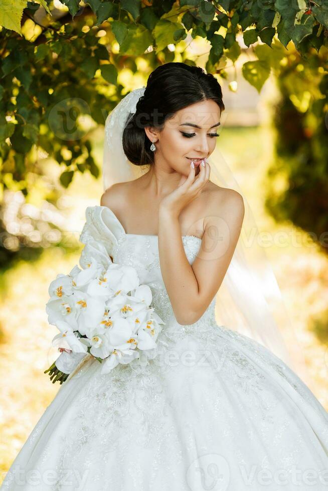 Warm sunny portrait of a happy bride with a bouquet of orchids in her hands, outdoors and at sunset. Warm summer time. A long veil, a luxurious white dress with a train. photo