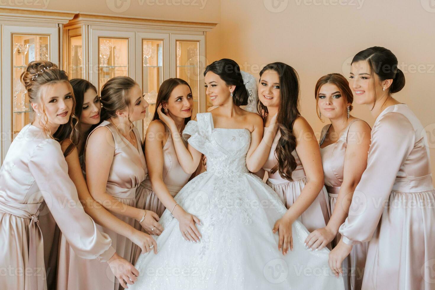 Stylish beautiful bridesmaids in matching silk dresses helping gorgeous brunette bride in white dress get ready for wedding, morning preparations, woman putting on dress photo