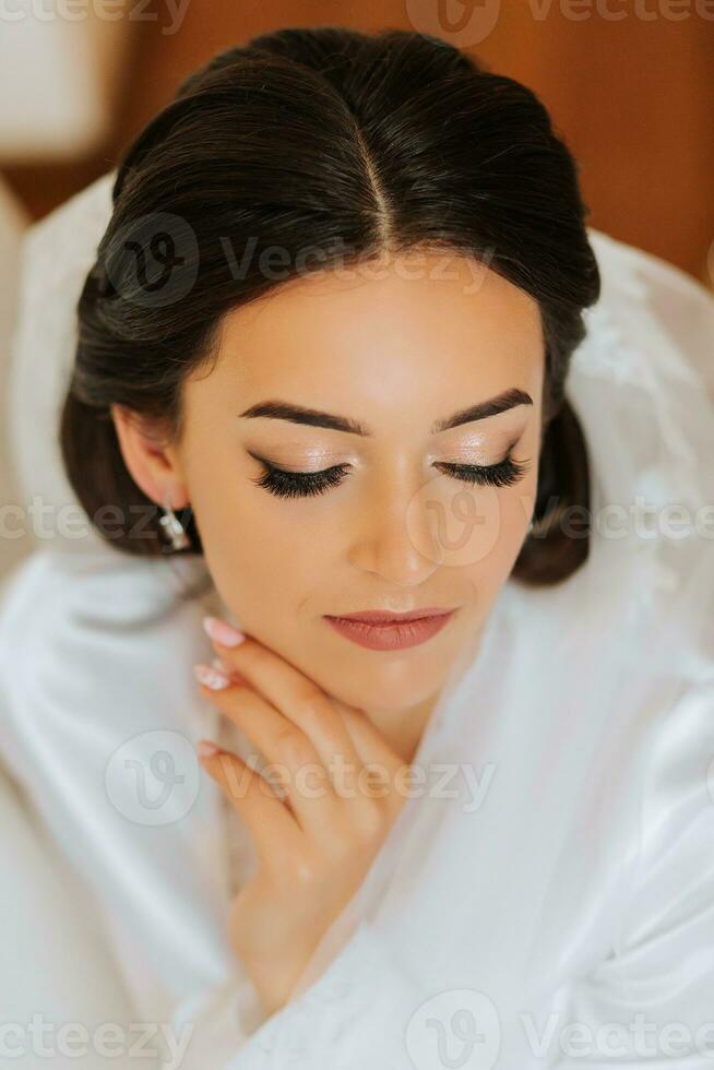 happy brunette bride in white satin gown with long veil professional makeup and hair. The bride is sitting in the room on the floor near the mirror. Wedding dress on a mannequin photo