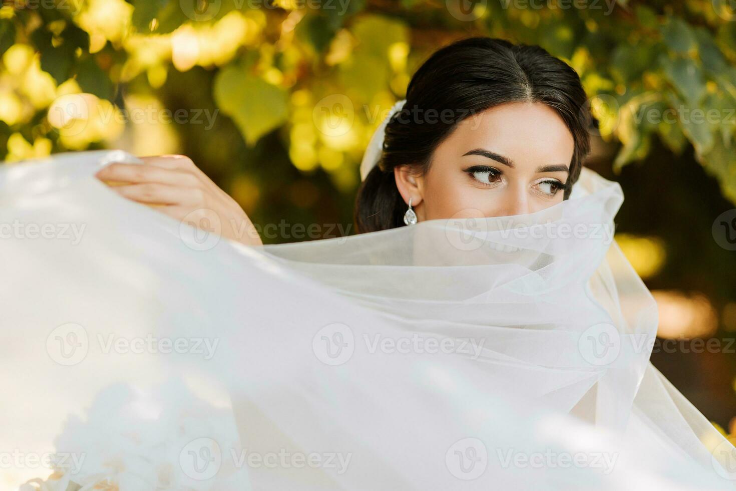 Warm sunny portrait of a happy bride with a bouquet of orchids in her hands, outdoors and at sunset. Warm summer time. A long veil, a luxurious white dress with a train. photo