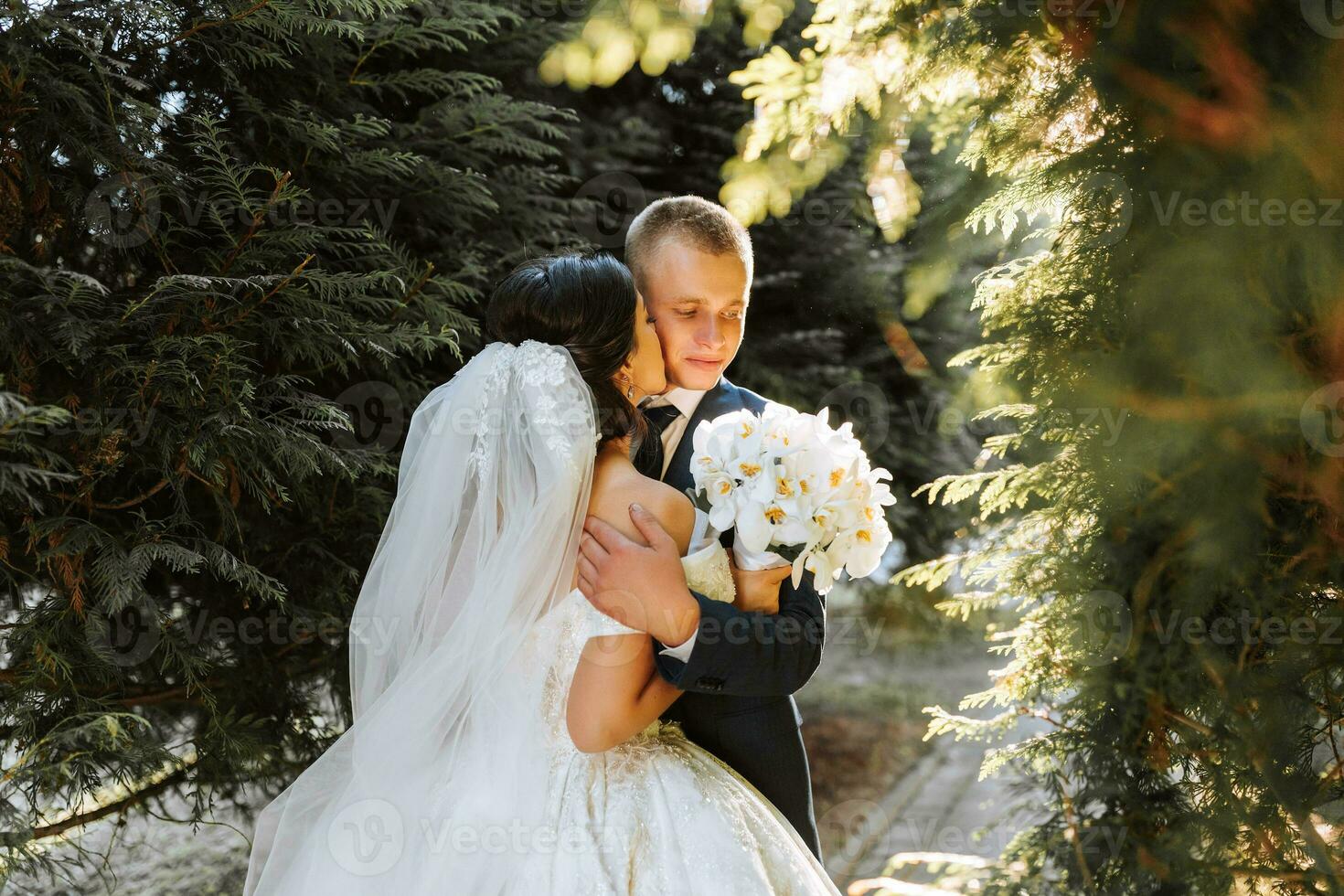 Happy bride and groom are walking in the park after the wedding ceremony. Long veil. Chic white dress with open shoulders. A bouquet of orchids. During sunset photo