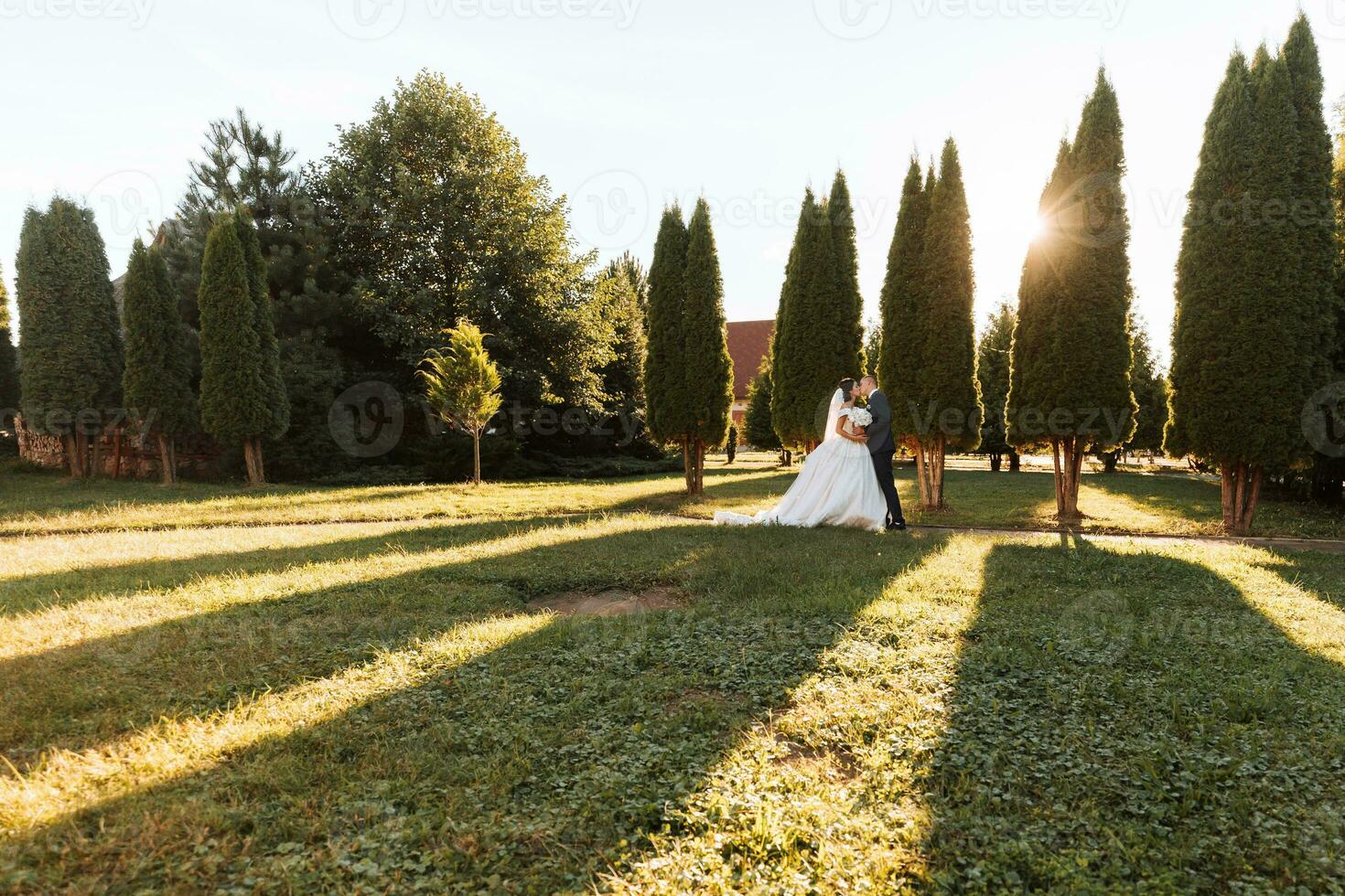 Happy bride and groom are walking in the park after the wedding ceremony. Long veil. Chic white dress with open shoulders. A bouquet of orchids. During sunset photo