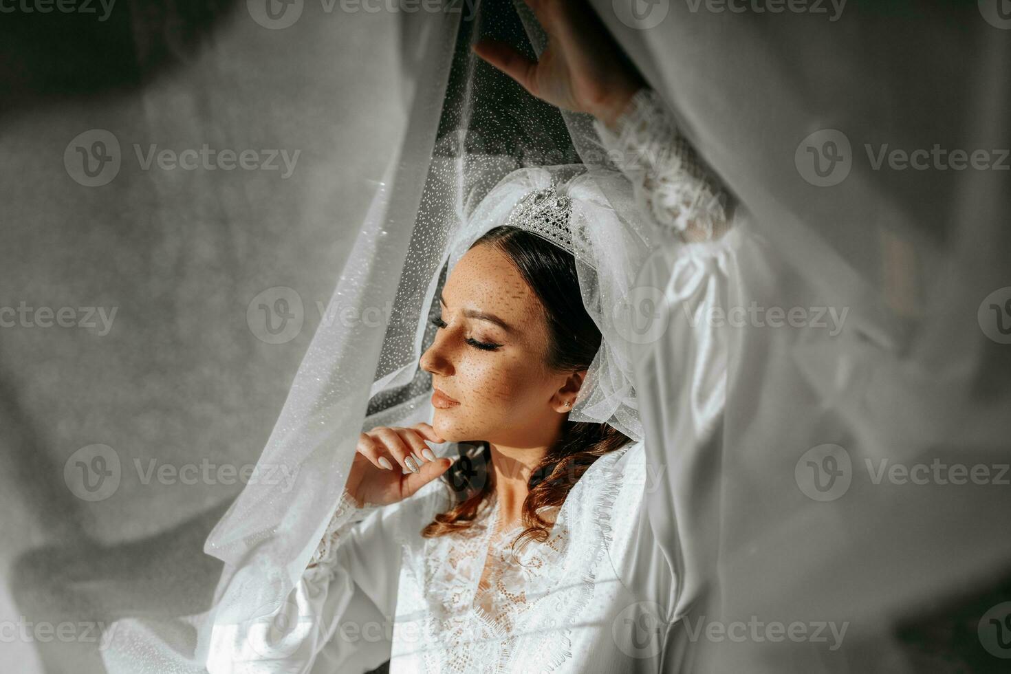 Beautiful bride with long dark curly hair in a long white robe in the morning. The bride is preparing for the wedding. Professional make-up and hair. A crown on the head and a long veil of the bride photo