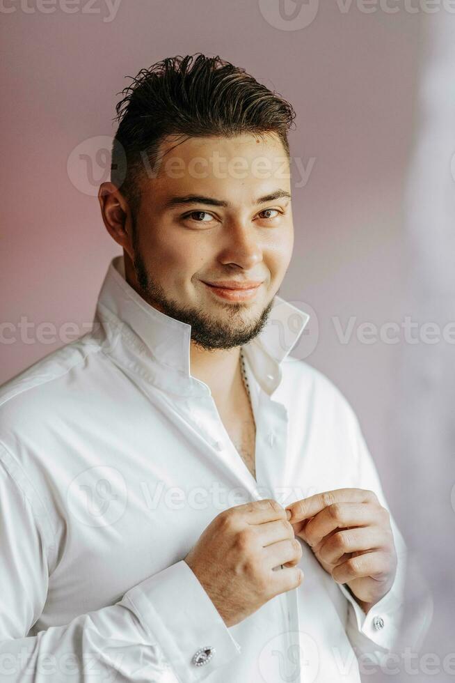 Close-up of a stylish man with a beard buttoning his shirt. The groom is preparing for the wedding. The man wears a white shirt. Stylish groom photo