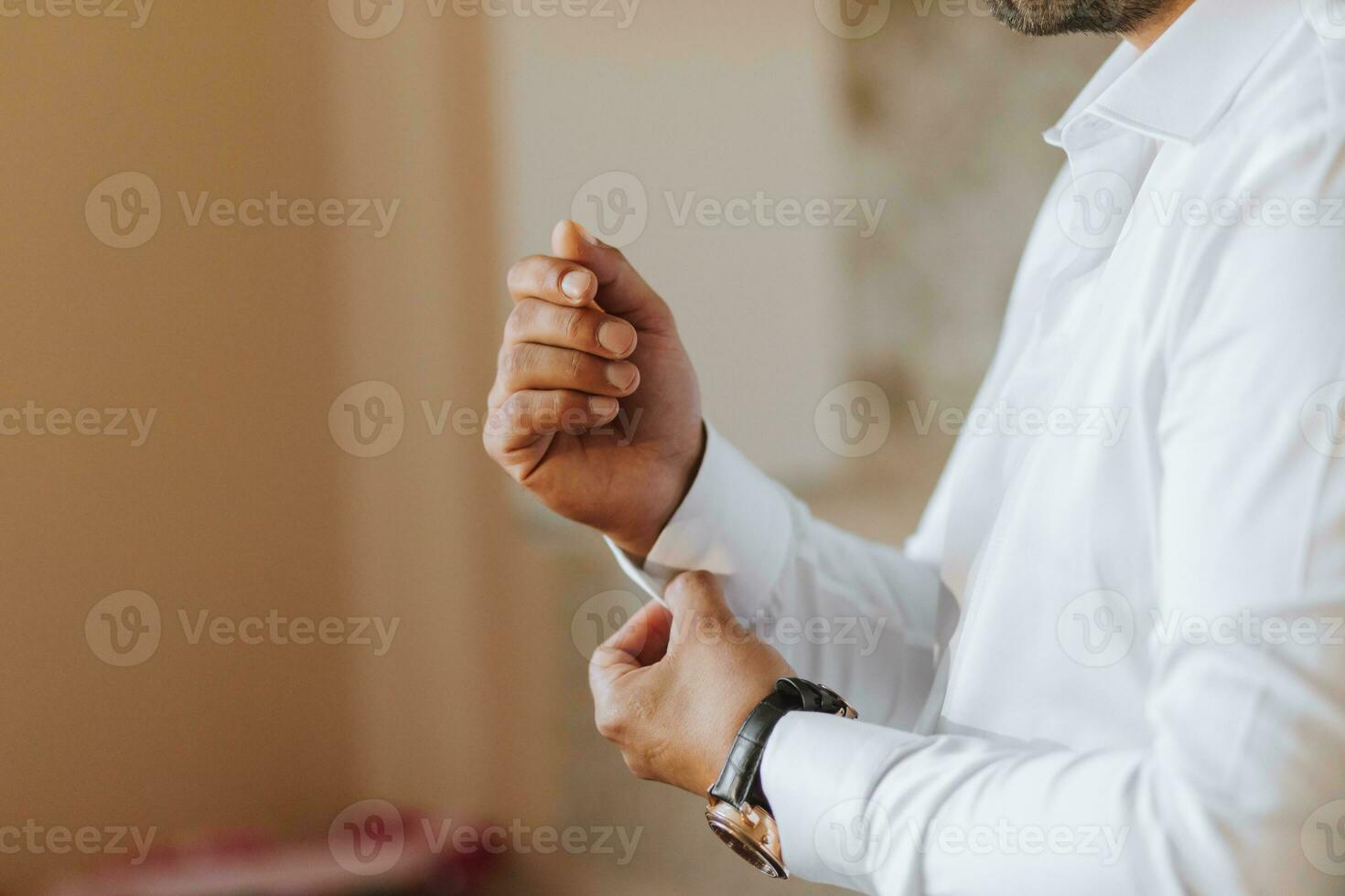 A young groom is buttoning up a white shirt while looking at the camera. The man is wearing a shirt. The groom is preparing for the wedding. photo