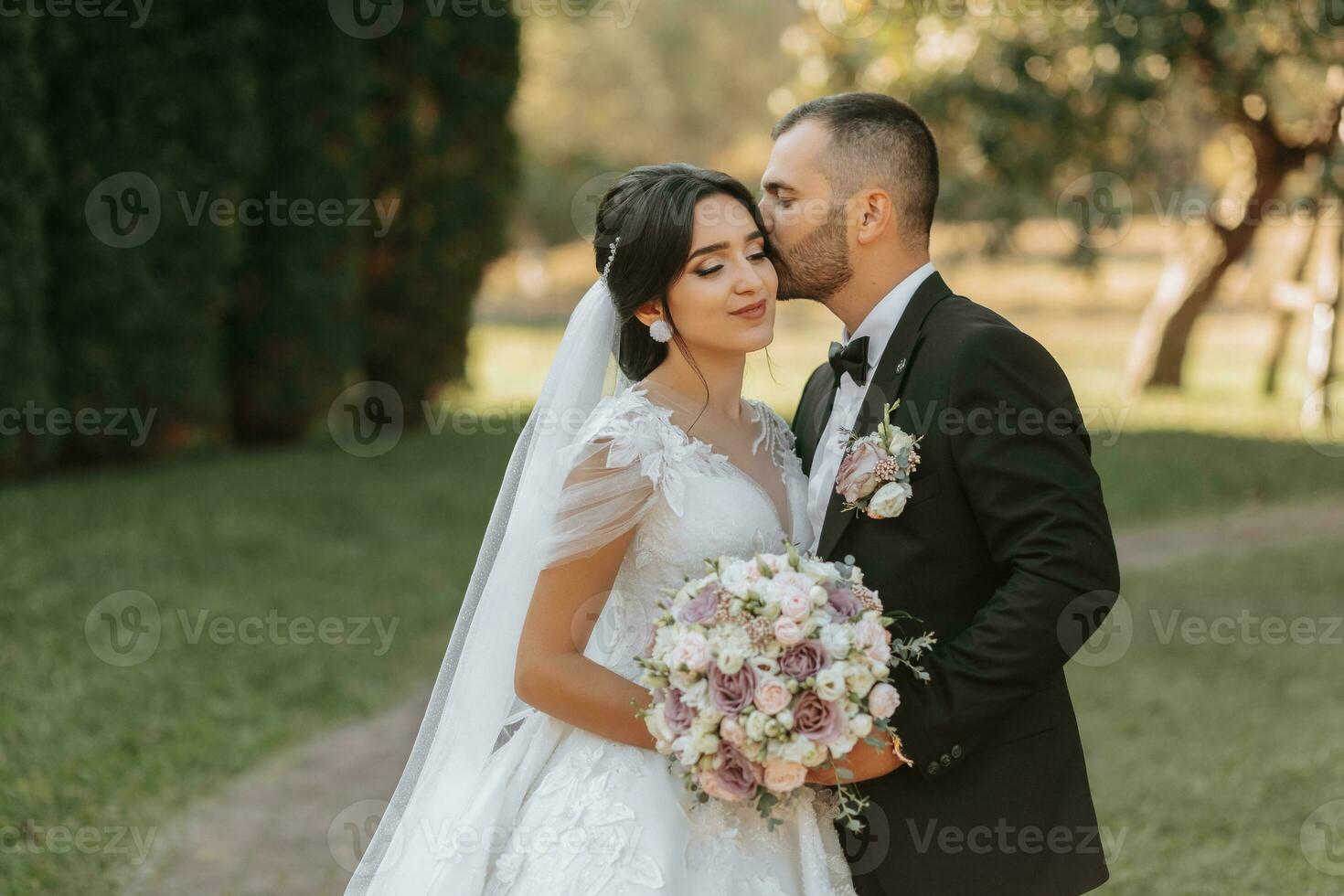 portrait of the bride and groom kissing against the background of the autumn sun of green trees in the garden or park. An incredibly beautiful bride. Stylish groom with a beard photo