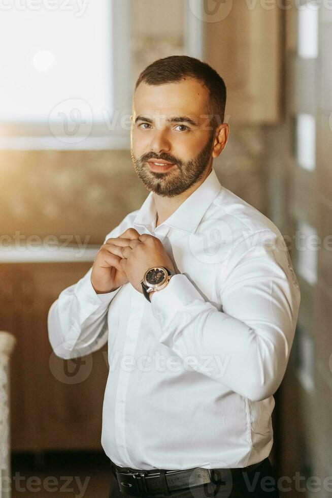 A young groom is buttoning up a white shirt while looking at the camera. The man is wearing a shirt. The groom is preparing for the wedding. photo