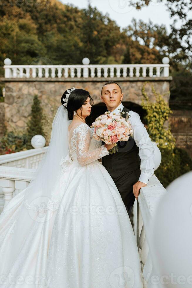 a beautiful bride in a chic wedding dress and an elegant groom stand on ancient steps in the park photo