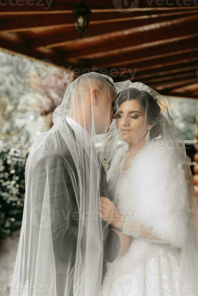 A man kisses a woman on the cheek. Bride and groom, happy, kissing under a veil near an authentic house photo