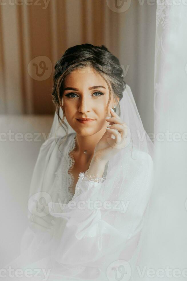 A stunning young bride poses by the window holding her veil in her hands. A beautiful woman of Caucasian appearance in an elegant white robe and luxurious hairstyle. Wedding makeup photo