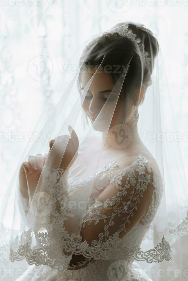 Female beauty. Cute woman at home. The bride poses for the photographer. A stylish woman wears a white dress. Professional make-up and hair. On the bride's head is a diadem and a long veil. photo
