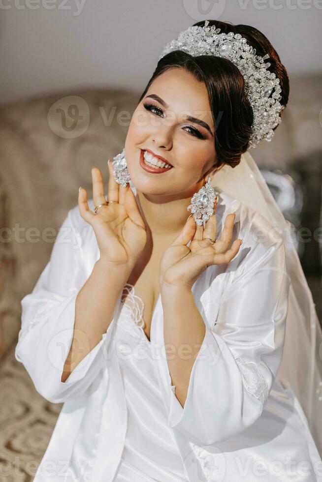 The bride in a white dress holds earrings in her hands. The bride shows off her earrings, dresses and prepares for the wedding. Professional wedding hairstyle and tiara on the head photo