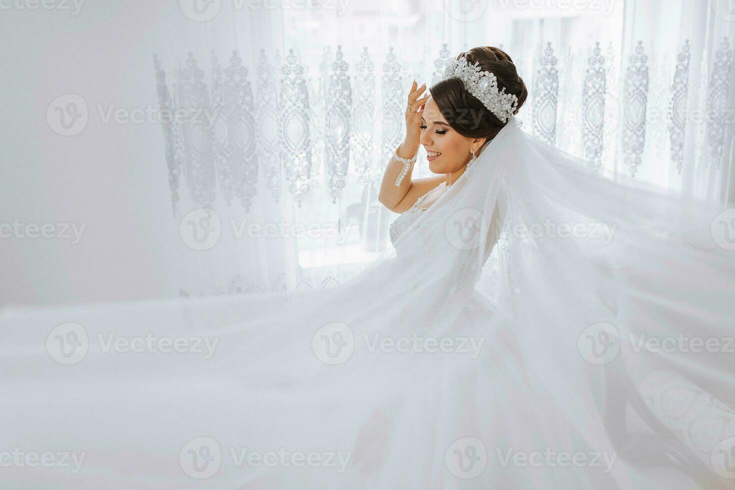 retrato de un novia con un hermosa peinado y tiara en su cabeza y natural maquillaje posando en un blanco túnica en su habitación. el belleza de el muchacha. un largo blanco velo foto