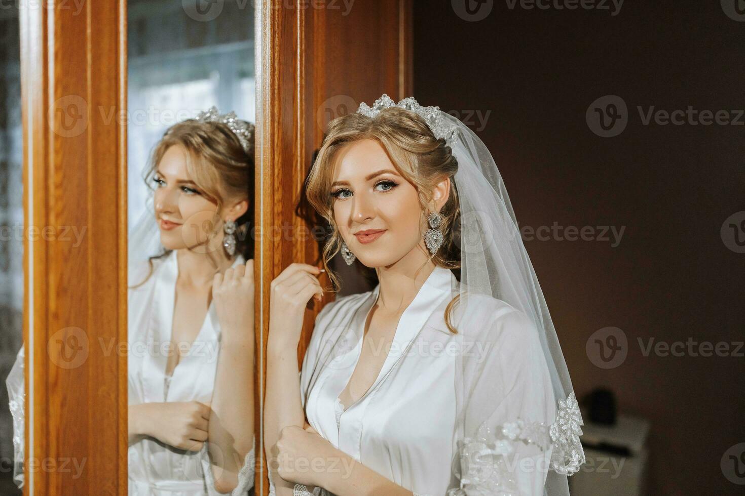 Preparation for the wedding. Beautiful young bride in a white robe in the room. A luxurious model looks at her reflection in the mirror with a tiara on her head. Morning of the bride photo