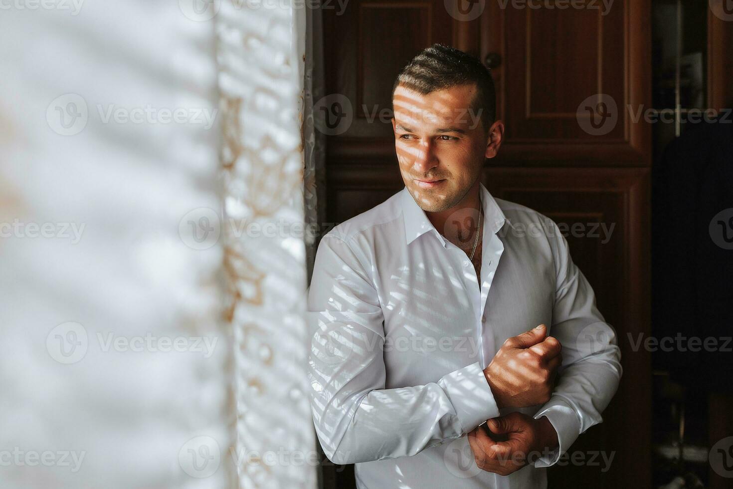 Handsome man putting on shirt standing near window at his room in morning. Preparing for some event or new workday. New opportunities, dating, wedding day or getting ready for job interview concept. photo