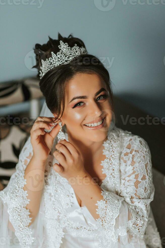 Beautiful young bride wearing earrings before wedding ceremony at home. The bride in a white robe puts on earrings in her room in the morning photo