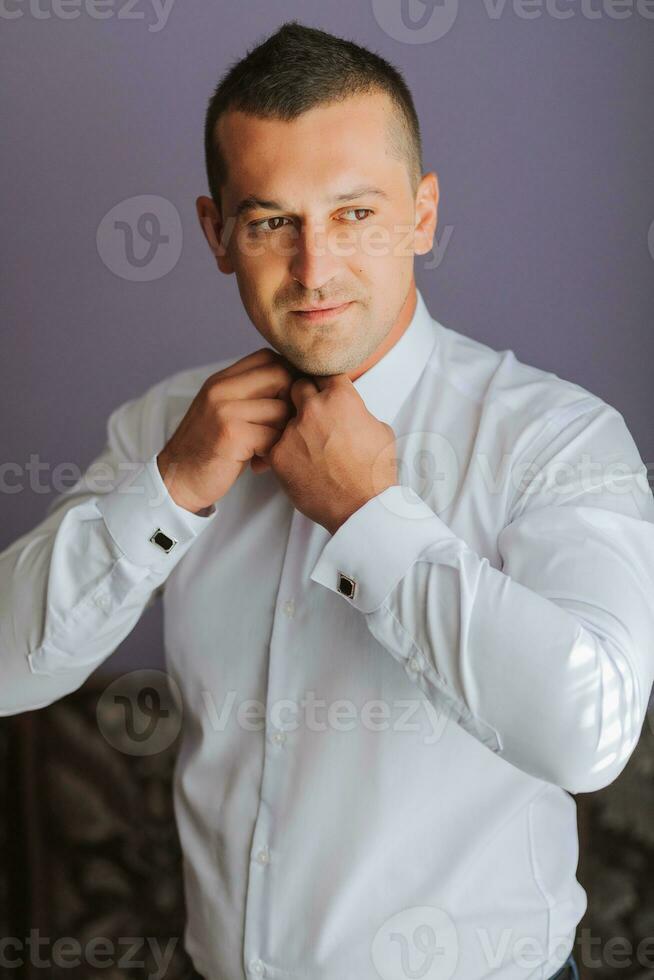 Handsome man putting on shirt standing near window at his room in morning. Preparing for some event or new workday. New opportunities, dating, wedding day or getting ready for job interview concept. photo