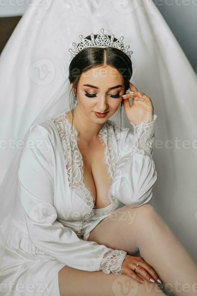 un increíblemente hermosa novia en el Mañana en un satín túnica con un tiara en su cabeza poses siguiente a su Boda vestir foto