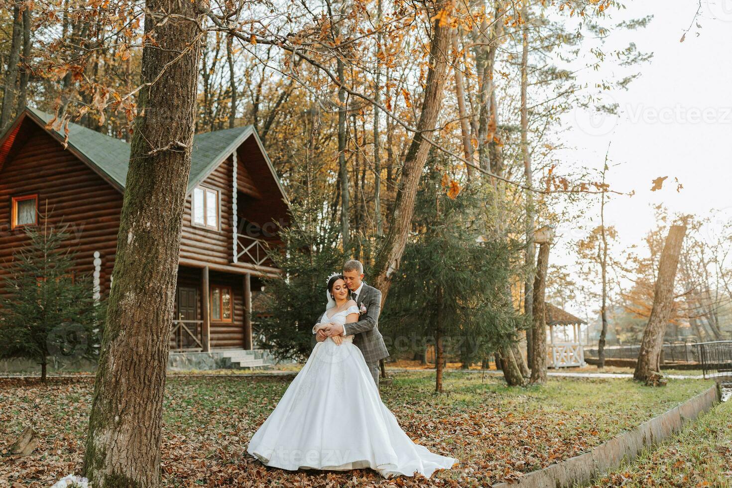 A fabulous romantic newlywed couple is hugging at sunset in an autumn park with leaves from trees on the grass. Wide angle photo