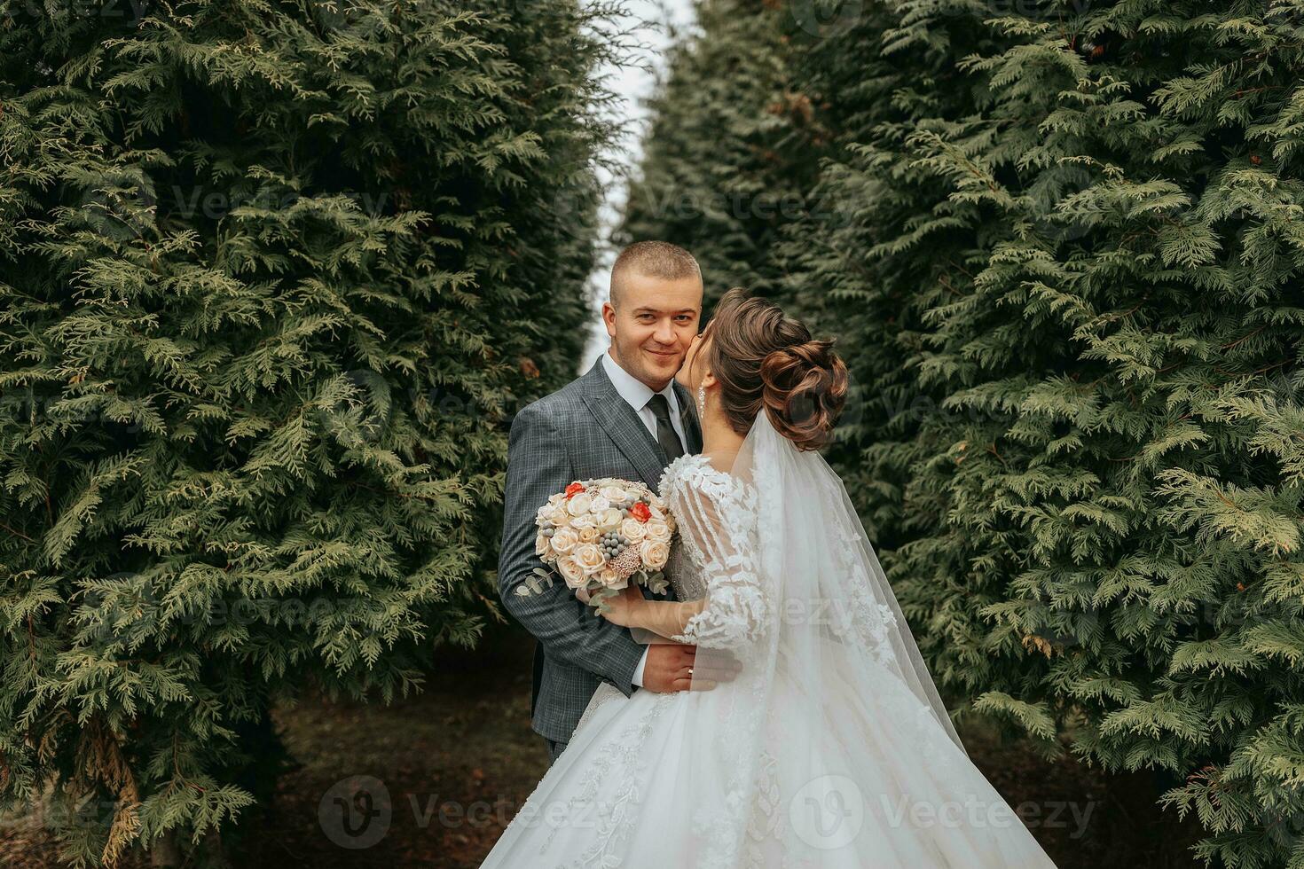 Stylish bride and groom tenderly embrace and kiss. Beautiful bride with modern bouquet embracing stylish groom in autumn park. Sensual romantic moment. Wedding. Stylish and beautiful. photo