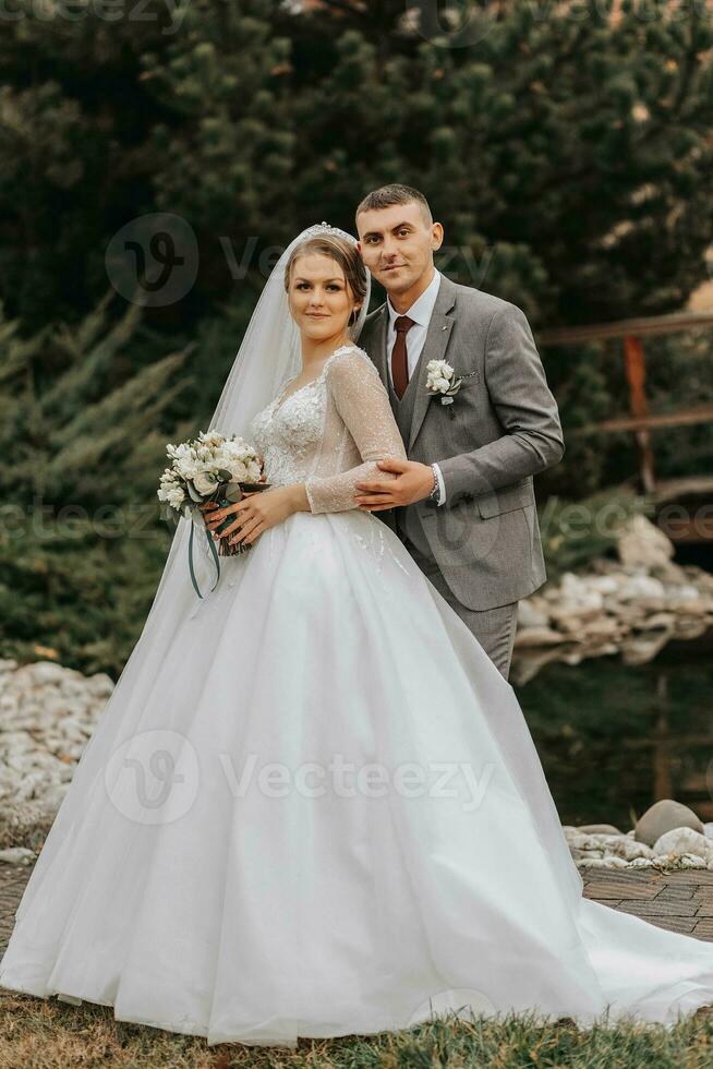 Stylish bride and groom tenderly embrace and kiss. Beautiful bride with modern bouquet embracing stylish groom in autumn park. Sensual romantic moment. Wedding. Stylish and beautiful. photo