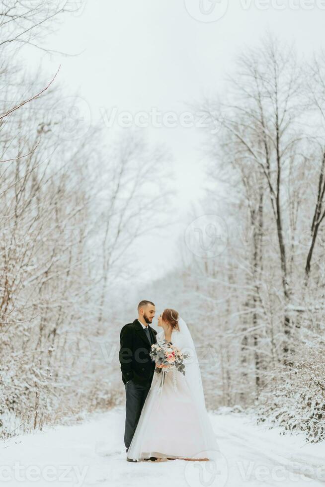 Beautiful wedding couple standing in winter snowy forest, woman in white dress and mink fur coat, bearded man in black coat photo