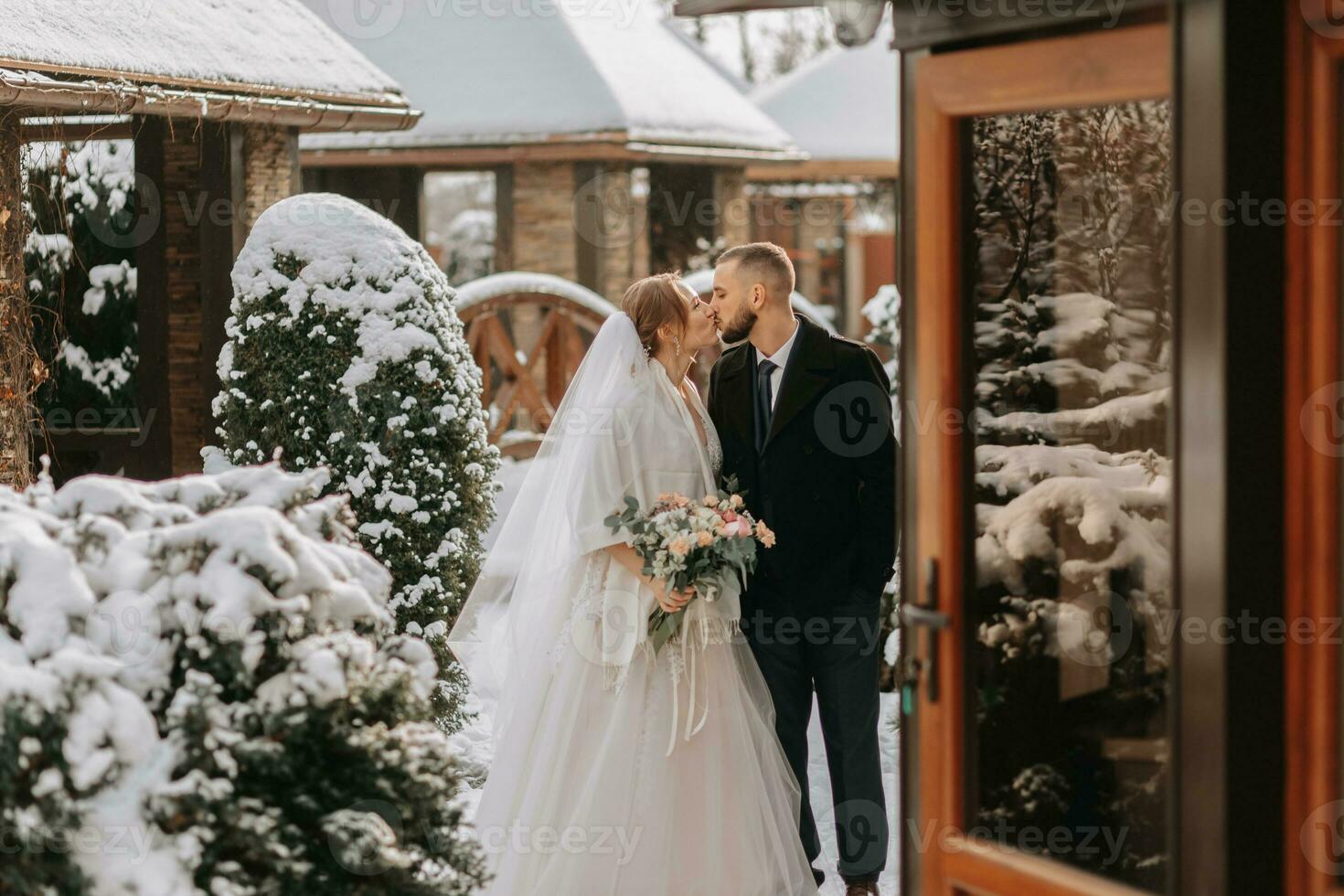 Beautiful and happy bride and groom are kissing between snow-covered trees. Bride and groom in the winter park. Bride with a bouquet of flowers in a wedding dress and poncho. Groom in a black coat. photo