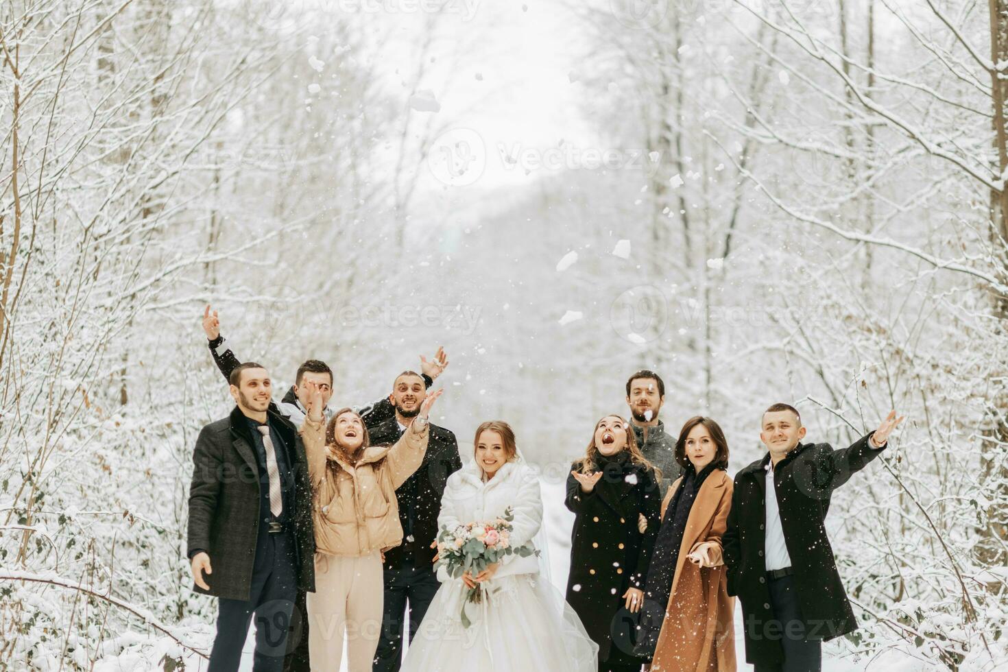 Happy newlyweds and their friends in winter coats stand in a snow-covered forest and throw snow with their hands photo