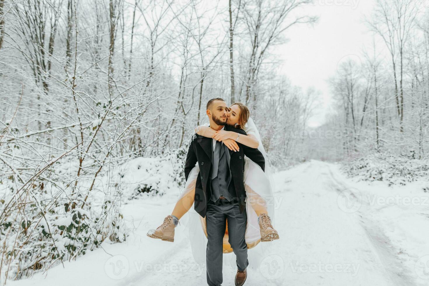 Beautiful wedding couple walking in winter snowy forest, woman in white dress and mink fur coat, bearded man in black coat. The groom carries the bride in his arms, a funny photo