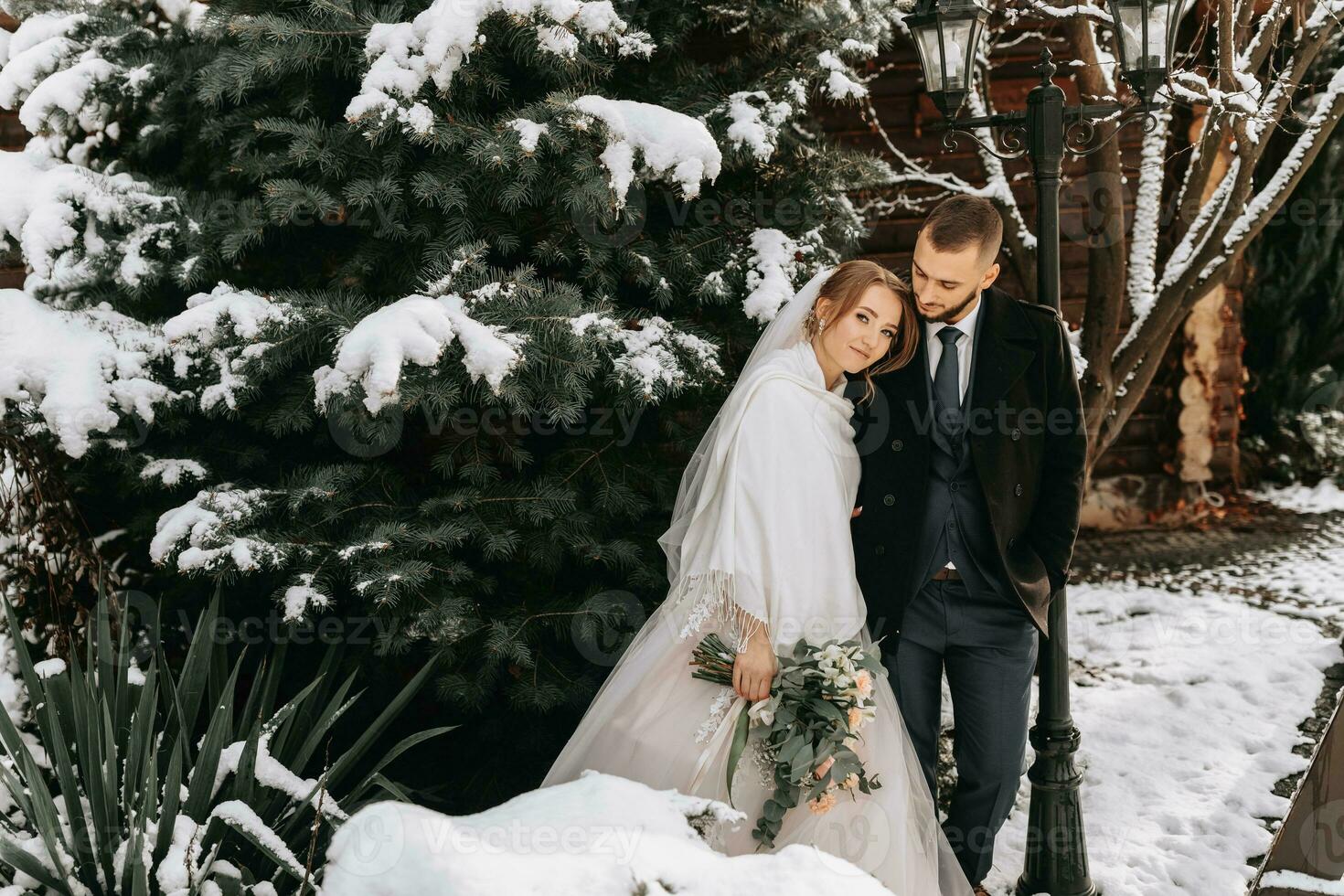 Happy bride and groom hug near snowy Christmas trees. Groom and bride in the winter park. Bride with a bouquet of flowers in a wedding dress and poncho. Groom in a black coat. photo