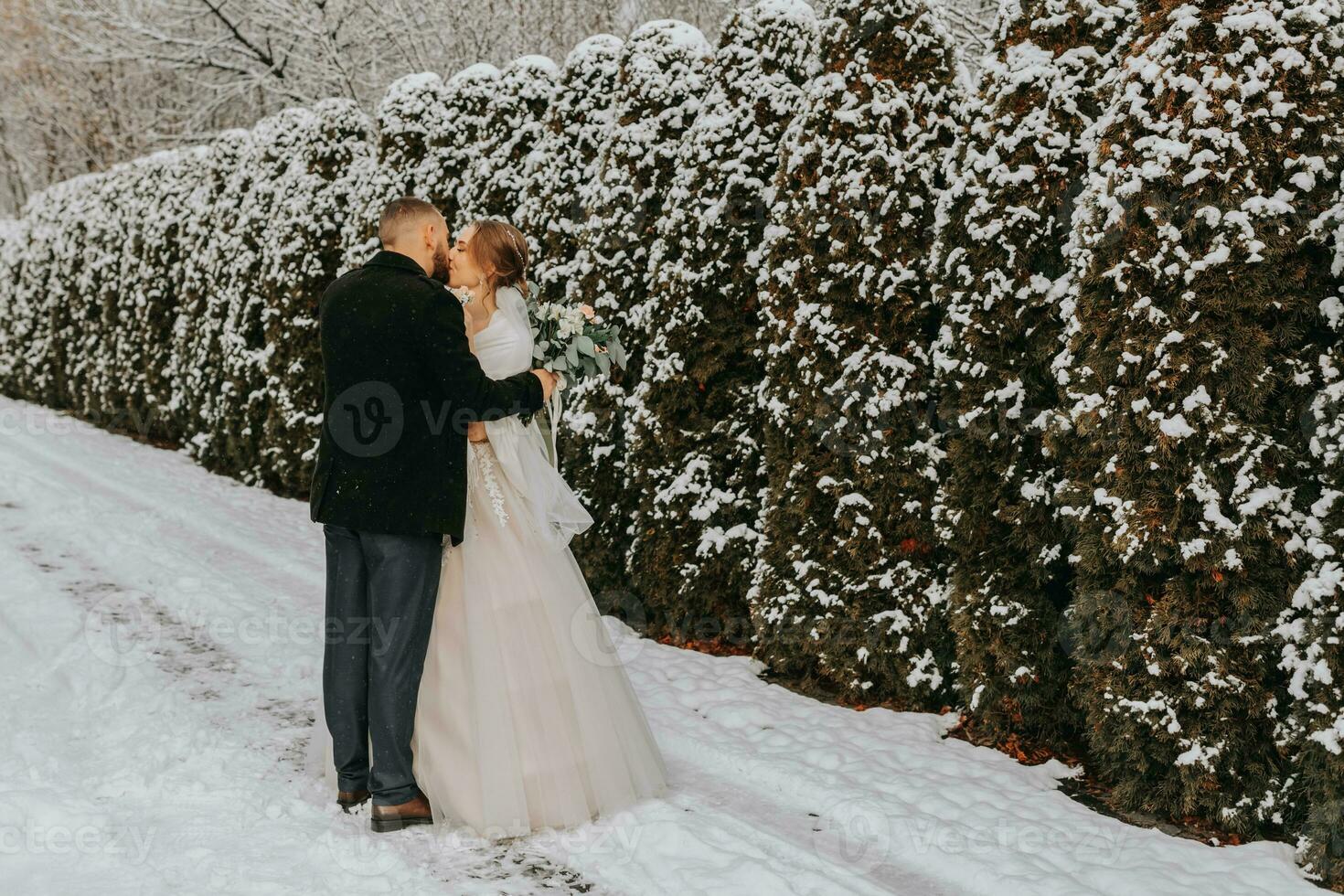 Sensitive portrait of happy newlyweds. The groom hugs and kisses the bride in the winter park. The bride in a wedding dress and poncho. The groom is dressed in a black coat photo