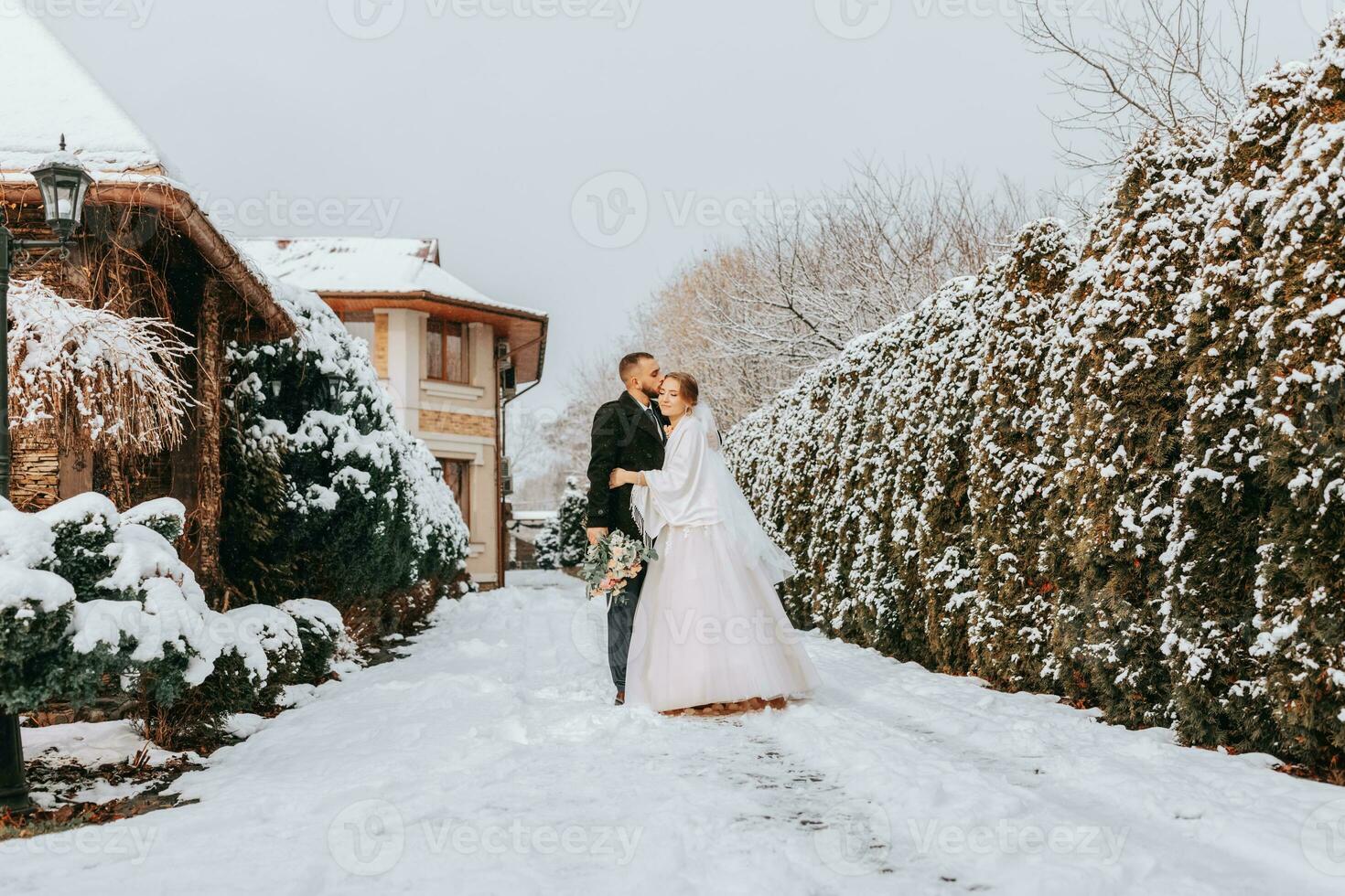 Sensitive portrait of happy newlyweds. The groom hugs and kisses the bride in the winter park. The bride in a wedding dress and poncho. The groom is dressed in a black coat. Wide angle. Free space photo